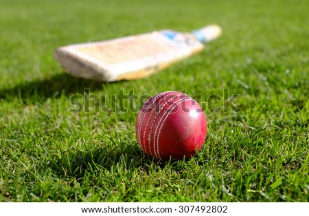 Red cricket ball on the grass with bat in the background