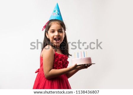 Cute adorable little Indian/asian small girl celebrating birthday while holding strawberry cake and blowing candles at table or standing isolated over white or red background