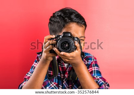 10 year old indian boy holding digital camera or DSLR camera, posing like a professional photographer, young photographer, kid photographer, child photographer, portrait, closeup, red background