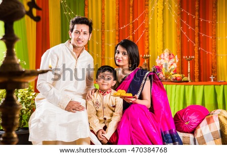 smart looking indian young couple in traditional wear sitting on sofa with boy child and ganesh idol or ganpati bappa in the background on ganesh festival or ganapati festival or utsav day