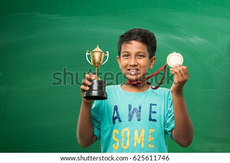 indian smart boy or school kids holding gold medal and trophy cup over green chalkboard background
