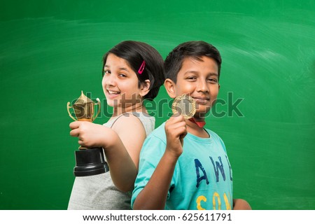 indian smart girl and boy or school kids holding gold medal and trophy cup over green chalkboard background