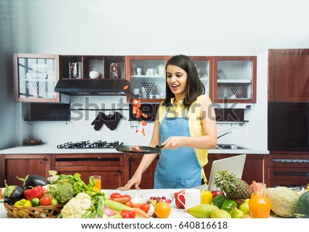 Attractive young Indian girl wearing apron in kitchen and tossing vegetables in frying pan, with table full of fruits and vegetables and computer