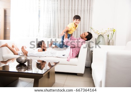 Happy Indian father playing with son while lying on sofa in living room at home. Asian Young man playing with small boy. Flying child enjoying playing with dad while sister watching