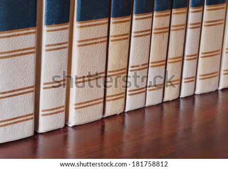 Close up of several volumes of a set of once white old books on a wooden shelf. The books are in varying thickness and shades of discoloration. The books are reflected in the soft sheen of the wood