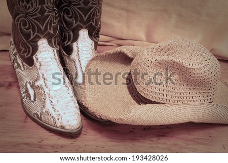 Detail of a pair of black leather with python skin cowboy boots and floppy straw hat on rustic wooden table of cedar. There is a soft golden backdrop. Vignette is added for a vintage atmosphere.