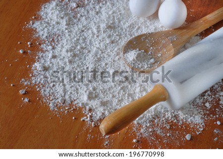 Image of baking elements on a wooden surface as viewed from above. Overhead perspective of flour tossed on table, eggs, wooden spoon and rolling pin.  Shades of brown and white. Horizontal composition