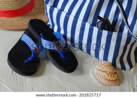 Summer beach scene of blue striped beach bag and flip flops on sand. There is a straw hat with a red band and a seashell beside the bag and shoes in the sand. Sunglasses are tucked into a pocket