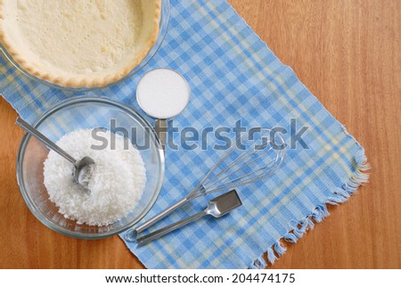 Baking elements and ingredients for a home made pie on a blue plaid placemat. Shot from directly above. Scene includes a pie shell, sugar, coconut and measuring spoons All items are on wood surface
