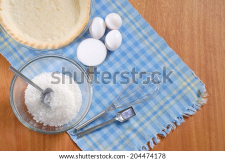 Baking elements and ingredients for a home made pie on a blue plaid placemat. Shot from directly above,, scene include a pie shell, eggs, sugar, coconut and measuring spoons on blue plaid placemat.