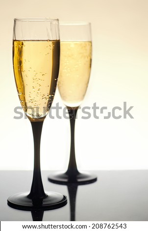 Two glasses of bubbly champagne or sparkling wine on dark grey reflective surface. Selective focus is on the near glass. Glasses have black bases and stems. Background fades from golden tan to white.