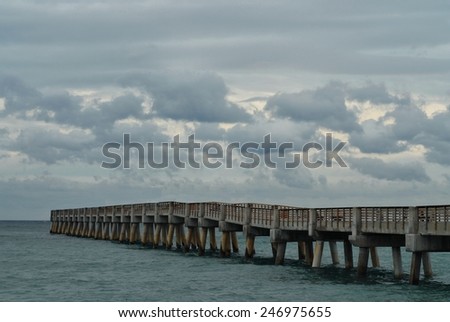 A long and wooden pier juts out into the ocean on a grey, overcast and cloudy day.  Horizontal image of Lake Worth Pier in Palm Beach County, Florida.  The pier would normally be crowded with people.
