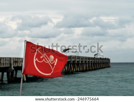 A red, no swimming caution flag is up on a stormy beachside location on the Atlantic ocean next to an empty pier.  The sky is overcast and cloudy and the flag shows how strongly the wind is blowing.