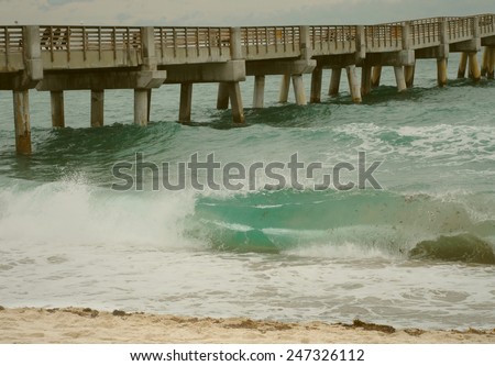 Sandy beach scene of waves coming ashore next to a wooden pier on a stormy day with instagram style filter used.  Seaweed is seen in the bluegreen wave, indicating a stormy day with rough conditions.