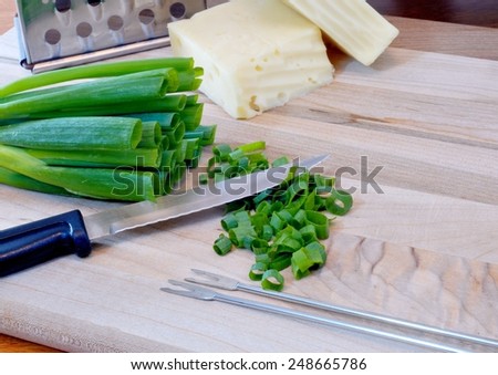 Ingredients and tools of cheese fondue include swiss and fontina cheeses, a cheese grater, green onions that have been chopped, fondue forks all on a cutting board with a fondue pot in the background