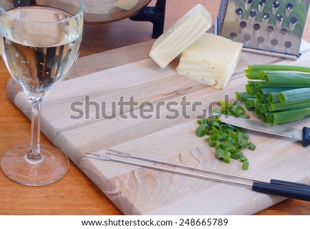 Ingredients & tools of cheese fondue include swiss and fontina cheeses, wine, a grater, green onions that have been chopped, & fondue forks all on a cutting board with a fondue pot in the background