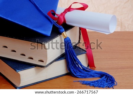 Detail of graduation themed items including blue cap and tassel with diploma tied up with a red ribbon. Both are on top of old books stacked on wooden table top in front of rough textured background