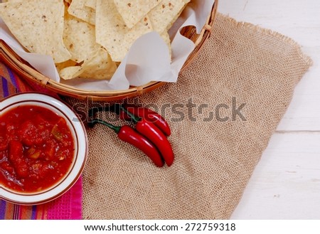 A basket of tortilla chips with a bowl of hot chunky salsa and some red peppers laying on a piece of burlap on a rustic wooden table. Cinco de Mayo message. Reddish orange is prominent color