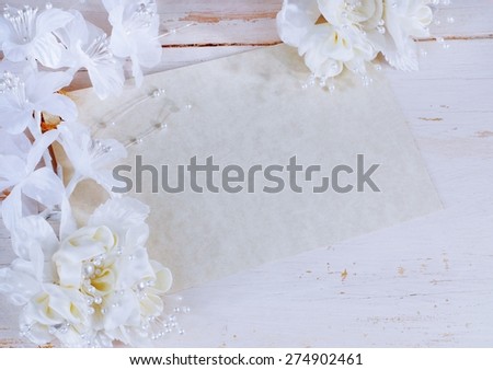 Horizontal image of blank mottled parchment paper card on rustic off-white wooden background. White and ivory colored silk flowers decorate the border of the card. Good for wedding or anniversary