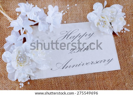 An ivory colored, parchment paper card that has white and cream silk flowers decorating the border. Happy 25th, or silver, anniversary is the message. The card is laying on a textured, burlap surface.