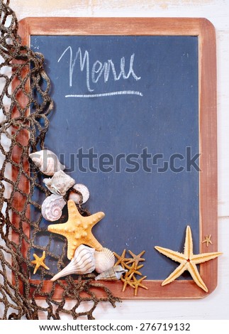 Seafood menu image of blackboard with fishnet and seashells and starfish gathered on left. The slate on the blackboard is rough and the surrounding brown frame is worn. Message is menu with copy space