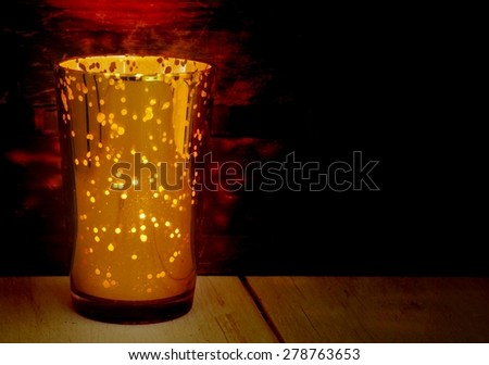 Low key image of a golden glass candle holder with many points where light can come through. Rustic wood textures below and behind the candle pick up the warm tones. Copy space on right side of image