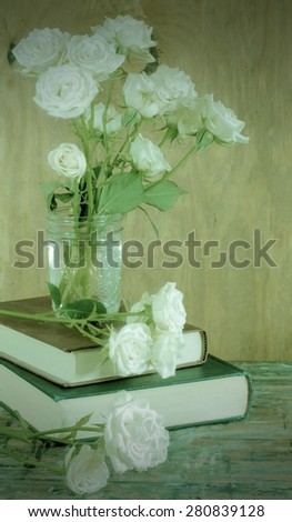 Tired, white miniature tea roses in a mason jar on a stack of dusty books. The table top is old wood with many paint stains. A rustic wooden wall is the background. Vintage instagram type filter used