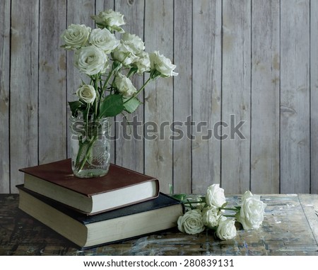 Tired, white miniature tea roses in a mason jar on a stack of dusty books. The table top is old wood with many paint stains. Wooden background with copy space. Low key side lighting and desaturation.