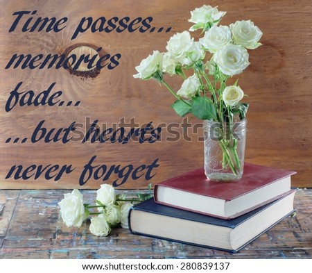Tired, white miniature tea roses in a mason jar on a stack of dusty books. The table top is old wood with many paint stains. A rustic wooden wall is the background. Memory message on left side