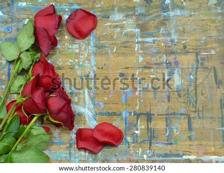 Wilted and faded red roses and green stems on a wood surface with multiple paint stains. Some fallen rose petals are scattered around. Concept of age or old love. Copy space on right.