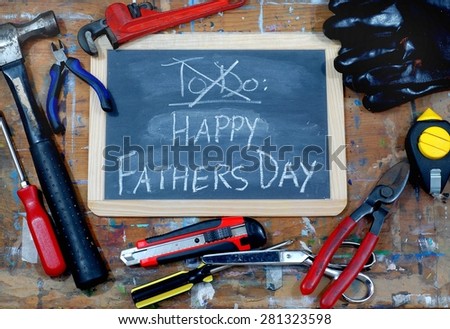 Dad\'s to do list has been crossed out and a Happy Fathers Day message has been written on a blackboard on paint stained table with various tools around, including a hammer, scissors and wrench.