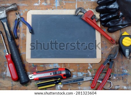 A blank blackboard on a paint stained table with various tools scattered around including a hammer, a wrench, snippers, a box cutter, a plumb bob, a screwdriver and scissors. Copy space on blackboard.