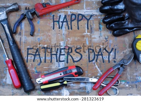 Happy Fathers Day message in the center of a paint stained work surface with tools scattered around. Tools include hammer, screwdrivers, wrench, snipper, plumb bob, gloves, box cutter and scissors.