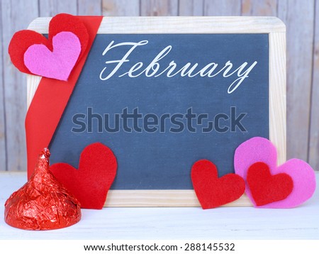 Month of the year for February with seasonally correct items placed around a blackboard with the month written across the top. There is a rustic wood background and copy space on the blackboard.