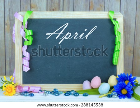 Month of the year for April with seasonally correct items placed around a blackboard with the month written across the top. There is a rustic wood background and copy space on the blackboard.
