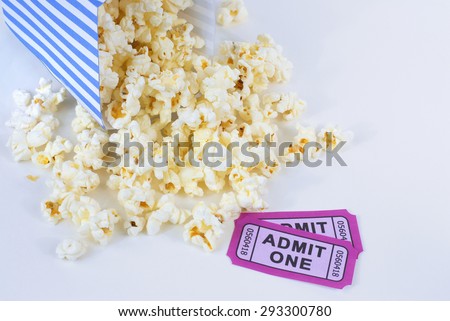 Summer movie theme with popcorn falling out of a tipped over blue and white striped bucket with two purple movie admission tickets. Popcorn is scattered about white background. Copy space.