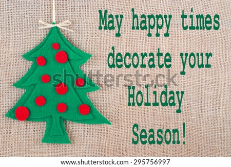 A Christmas tree shape cut out of green felt with round, red felt decorations, topped with a grass bow, is on a natural background. There is a seasonal message added. Rustic, homemade ambiance.