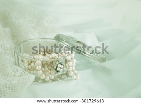 Antique pearls in a vintage crystal jewelry box with aged silver lid all on lace and cotton fabric. The lid is off the box and some pearls are falling out of the box. Copyspace. Vintage filter applied