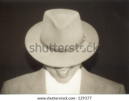 sepia toned portrait in soft focus of man grinning while wearing a hat. The man is looking down, so the brim of his fedora is covering his eyes. Isolated on black, he is wearing a suit jacket