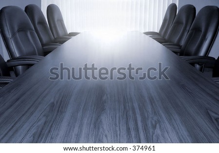 Empty boardroom table and chairs for at least six people in monotone grey/blue. Photo is taken looking towards a window with vertical blinds. Copy space upper in the bright window or lower on desk