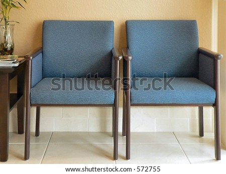 Two empty chairs in waiting or reception area. The chairs are upholstered in blue fabric. They are sitting on a tile floor in front of a gold colored wall. There is a side table with a plant and books