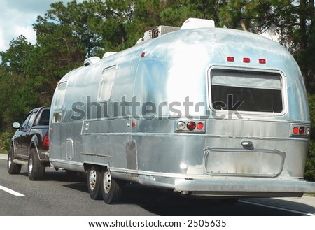 American vacation rounded retro style camper-trailer being towed down the highway by a pickup truck. The trailer is shiny silver and has a double axle. There is a rear window and proper lighting