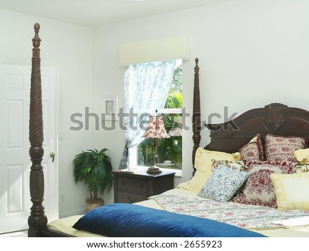 Sunshine streams through a window beside a four poster bed.  The bed is dressed with a mixture of country chic prints in blue, red and yellow. There is a night stand under the window next to a plant.