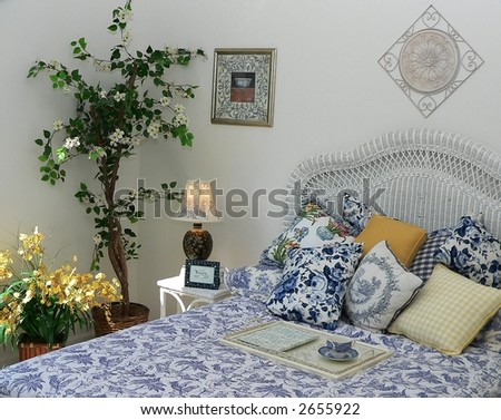 Pretty corner of a feminine styled bedroom includes silk plants, a white wicker headboard, blue toile bedspread and pillows. There are yellow accents with gingham pillow and a spray of forsythia.
