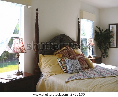 A comfortable, country chic four-poster bed is flanked by two, sunshine filled windows.  The nightstands have matching lamp.  The inviting bed is dressed in yellow with toile accents in red and blue