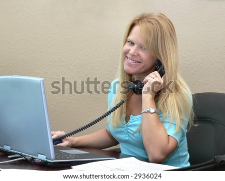 pretty blond woman with friendly smile for the viewer while on the telephone and at work on a laptop