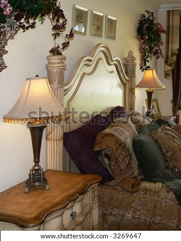 Bedroom scene of dressed bed and night stand. The décor is French stye and is heavy and ornate. The furniture is light in color and the tapestry bedding is in tones of gold, burgundy and green