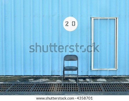 Folding metal chair sits empty beside door numbered with twenty in blue steel building. In front of the chair are steel storm drains. Only half the door is visible. The chair is directly below the 20.