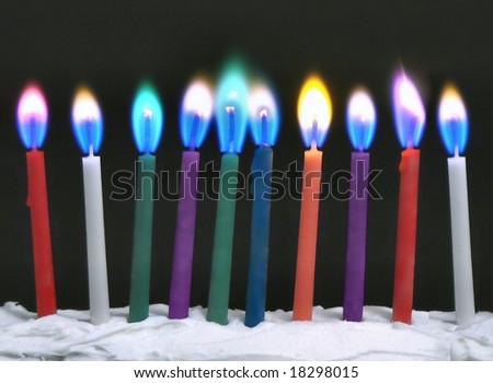 Birthday candles with colored flames on birthday cake with black background. The candles are red, white, green, purple, blue and orange. The flames are in shades of blue, green, pink and yellow.