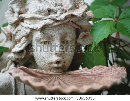 Detail of garden statue appearing to sip water from a shell - shallow depth of field. The statue depicts a fairy, or nymph with a beautiful, childish face. Her carved hair is tousled and messy.